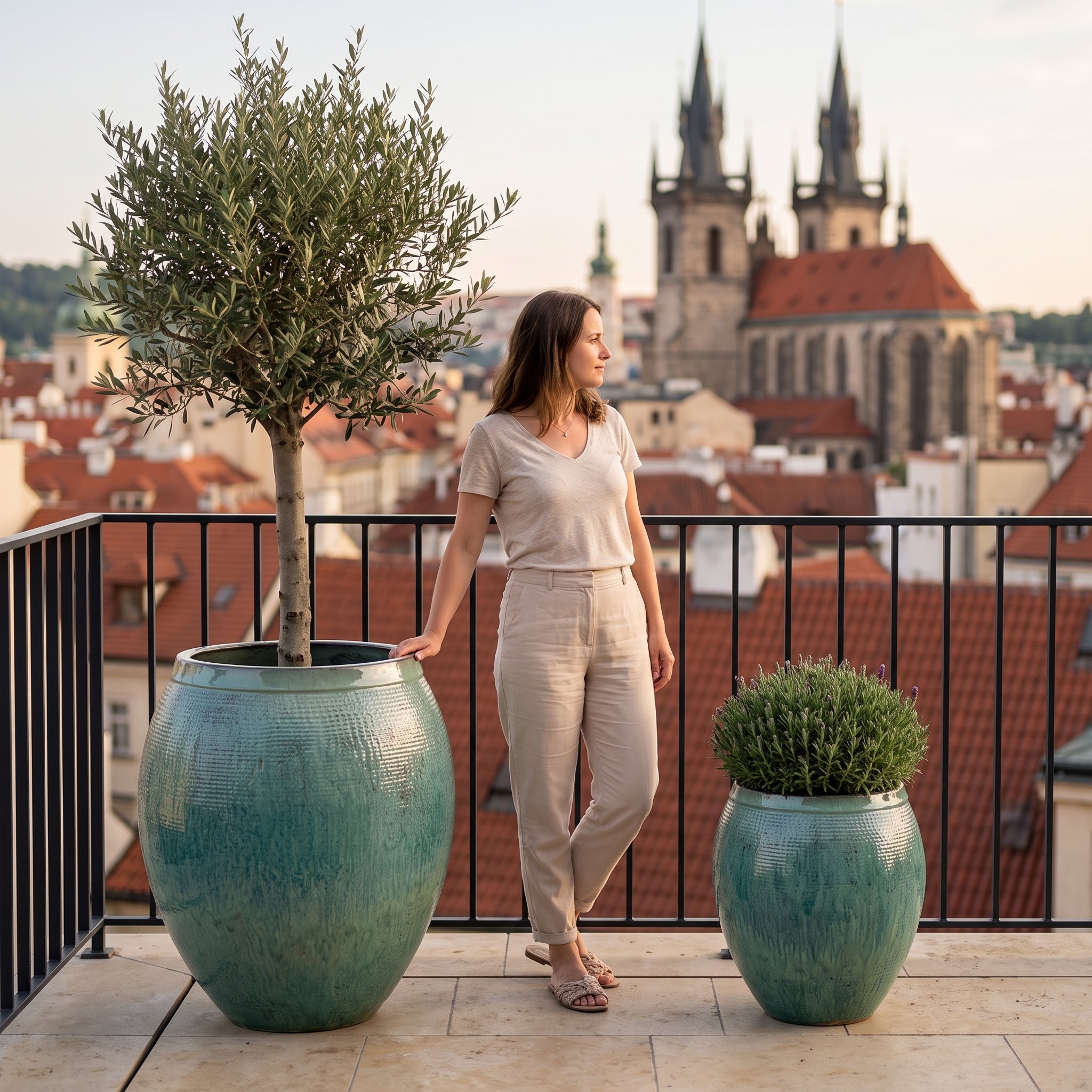 Velké ručně vyráběné terakotové květináče COCOON v oceánské modré barvě (velikost XXL s olivovníkem a XL s levandulí) na pražském balkonu. Žena stojící mezi nimi ukazuje jejich skutečnou velikost.