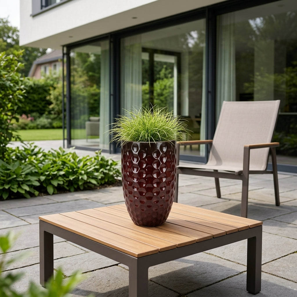 Keramický květináč MEROVEE - Cihlově červená - M (23 x 35 cm) osázený okrasnou trávou, designově umístěný na zahradním konferenčním stolku na terase moderního domu.